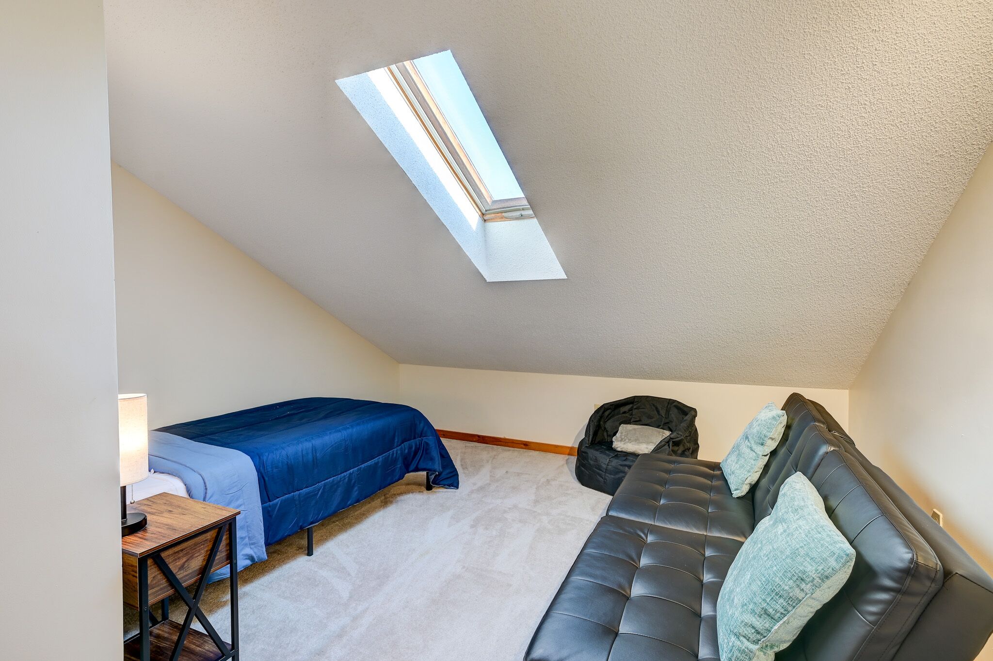 A bedroom with a sloped ceiling, a skylight, a bed with a blue bedspread, a black sofa with cushions, and a small table with a lamp.