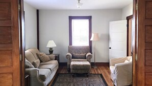 Una habitación con suelo de madera, un tapete estampado, un sofá, un sillón, una lámpara y una ventana con persianas.