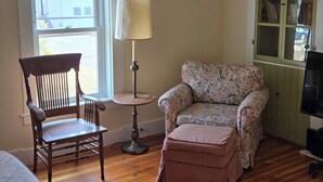 A room with a wooden chair, a small round table with a lamp, a floral armchair, a matching ottoman, a green cabinet with books, and a television. - Historic downtown apartment. Unscented products and sustainability practices. (Northampton)
