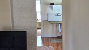 A room with a television on a desk, a white brick wall, wooden flooring, a patterned rug, and a kitchen area with a white cabinet and a radiator. - Historic downtown apartment. Unscented products and sustainability practices. (Northampton)