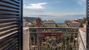 Un balcon doté d’une balustrade en métal, de plantes en pot et offrant une vue sur une ville côtière, avec des bâtiments et des montagnes visibles au loin.