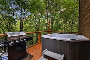 A deck with a Char-Broil grill, a hot tub, and a wooden railing overlooking a forest.
