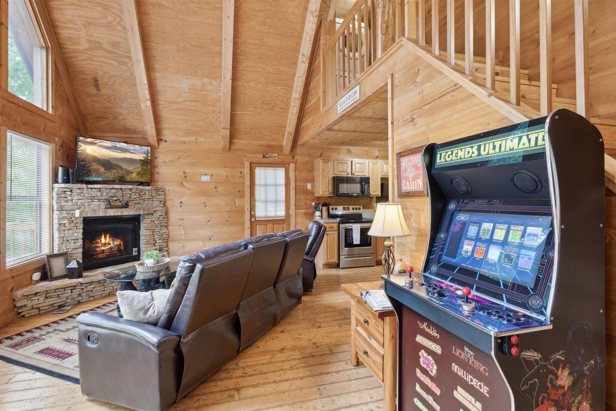 A cozy cabin interior with a stone fireplace, a large leather sofa, a kitchen area, and a gaming console.
