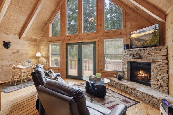 Cozy cabin with a high ceiling, leather sofa, and stone fireplace, perfect for relaxing and taking in the scenic forest views.