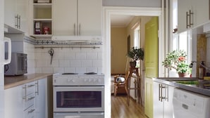 Een keuken met witte kasten, een fornuis, een magnetron en een vaatwasser. Er is een eethoek met een tafel en stoelen, en een plant op het raamkozijn.