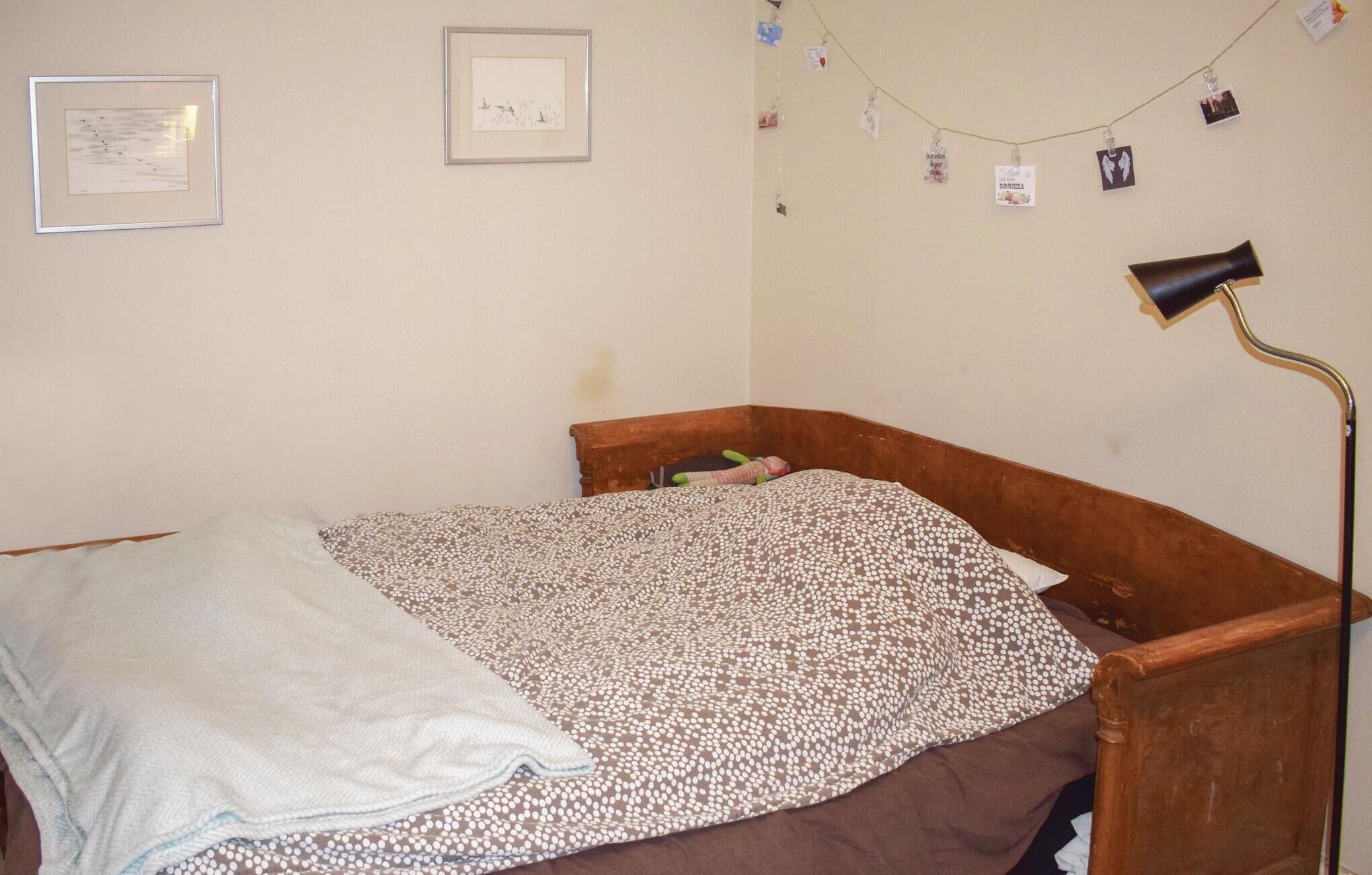 A bedroom with a wooden bed, a white blanket, a patterned bedspread, a desk lamp, and two framed pictures on the wall.