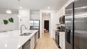 A modern kitchen with stainless steel appliances, a central island with a sink, and a view into the dining area. - NICE 15BR Home Near Disney | Groups Welcome (Kissimmee)
