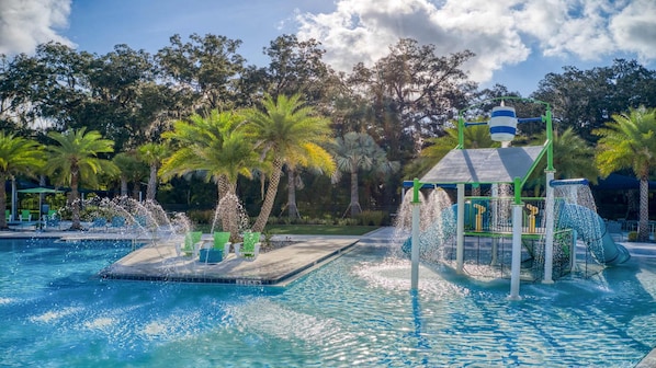 A swimming pool with water features, including fountains and a slide, surrounded by palm trees and a covered play area.