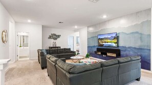 A modern living room with a sectional sofa, a coffee table, and a flat-screen TV mounted on a wall with a scenic landscape painting. - NICE 15BR Home Near Disney | Groups Welcome (Kissimmee)
