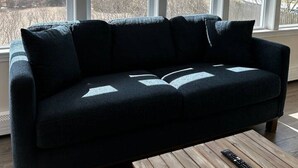 A dark blue sectional sofa with a remote control on a wooden coffee table in a room with large windows. - Cottage | 3 Min Walk to the Beach (Yarmouth)