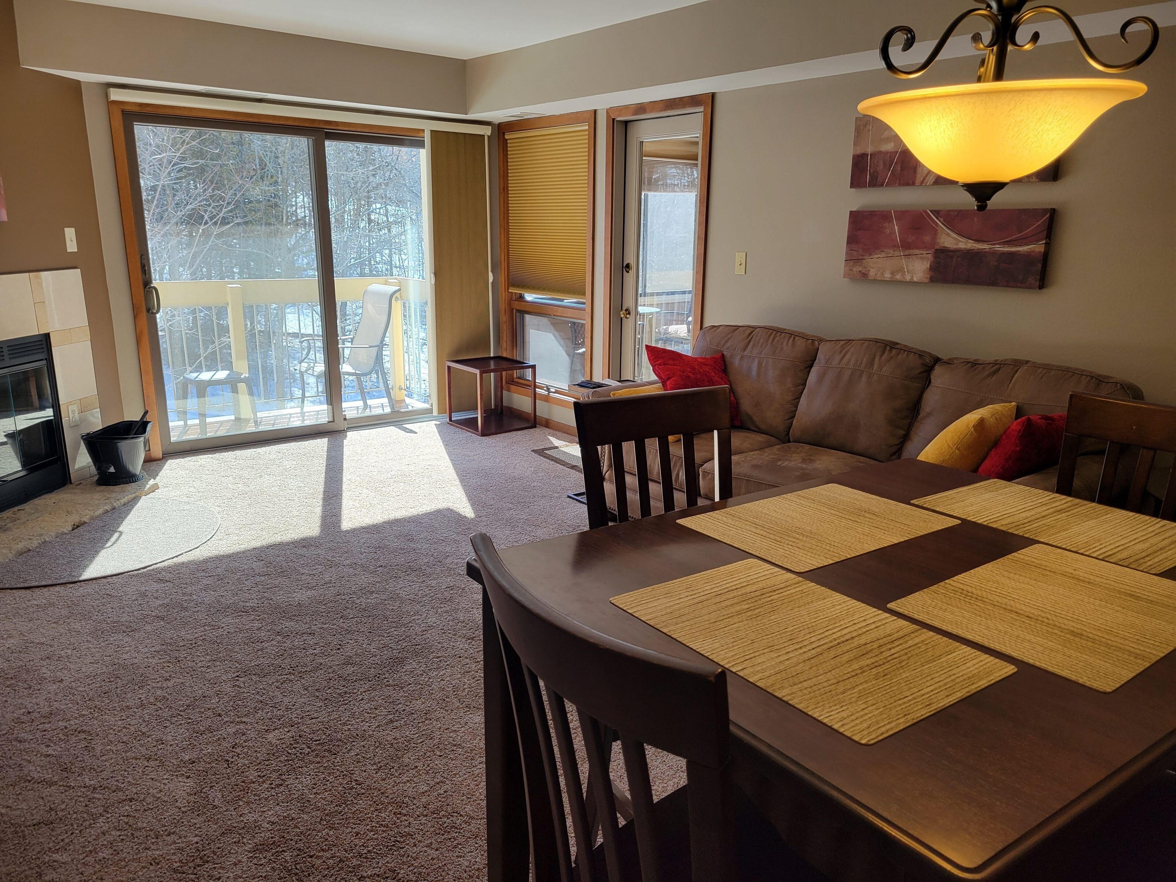 A living room with a brown leather sofa, a wooden dining table with placemats, a black chair, and a sliding glass door leading to a balcony.