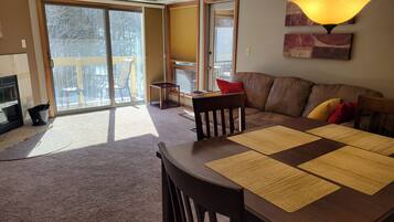 A living room with a brown leather sofa, a wooden dining table with placemats, a black chair, and a sliding glass door leading to a balcony.