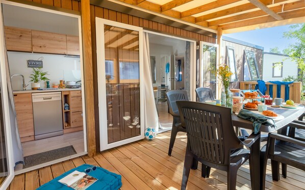 A wooden deck with a dining area, a kitchen, and sliding glass doors leading to an indoor space.