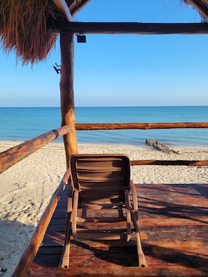 Eine Holzterrasse mit einem einzelnen Liegestuhl, der zum Strand hin zeigt.