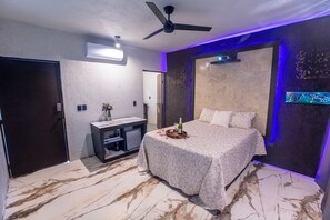A hotel room with a bed, a desk, an air conditioner, a ceiling fan, and a wall-mounted TV.
