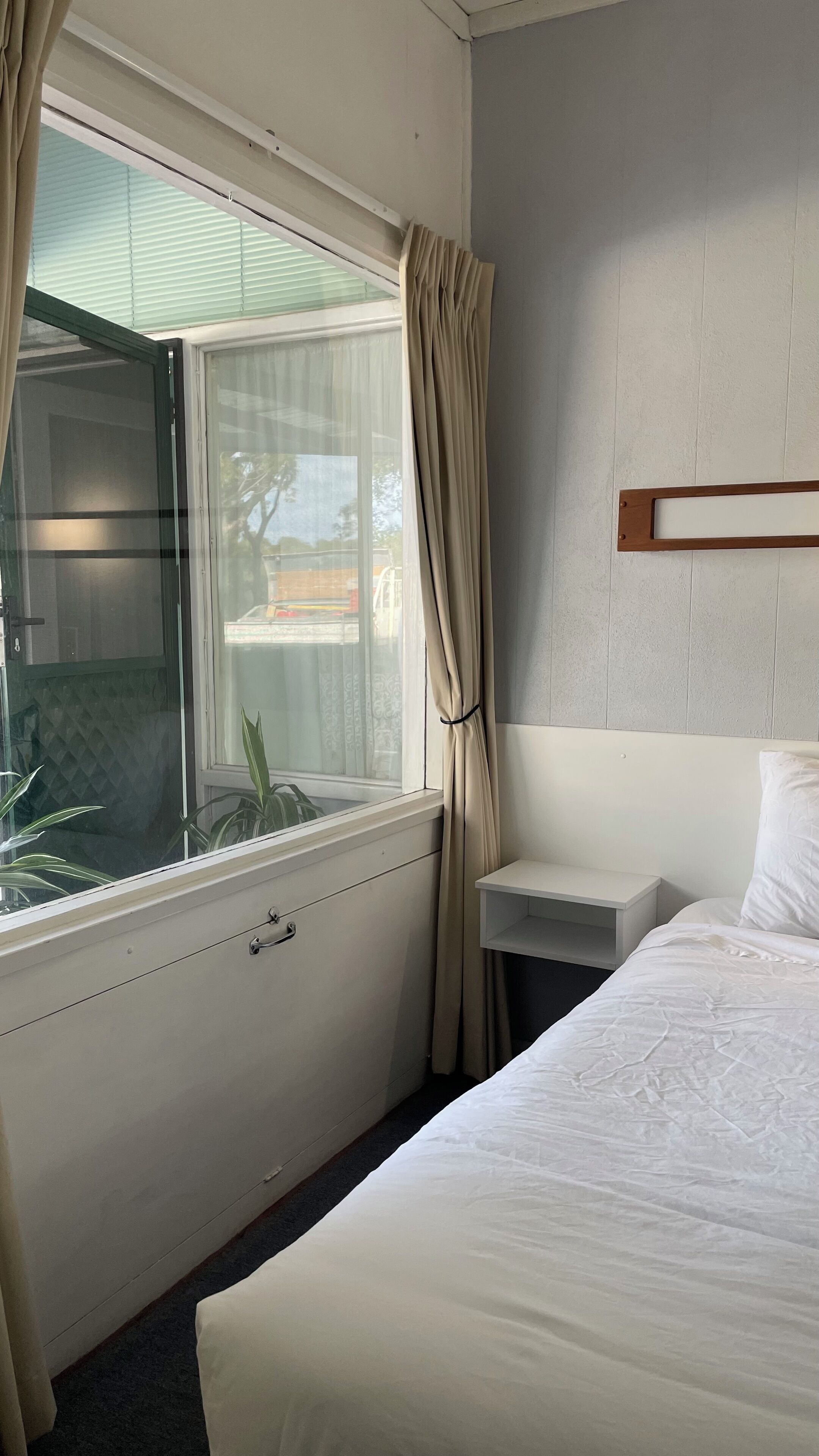 A hotel room with a bed, a nightstand, a window with curtains, and a view of the outside.