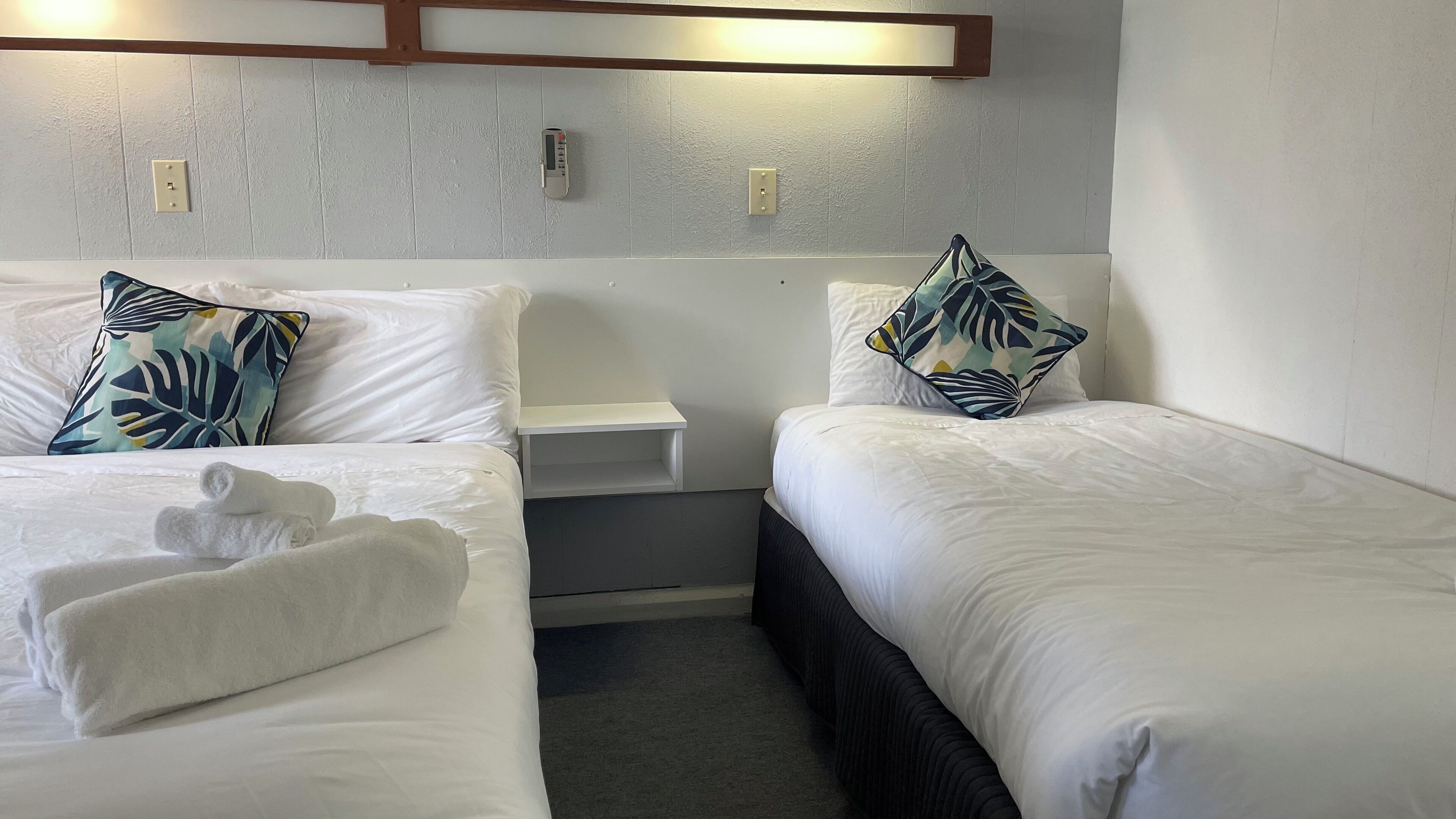 A hotel room with two beds, white bedding, a small bedside table, a nightstand, and a wall-mounted thermostat.