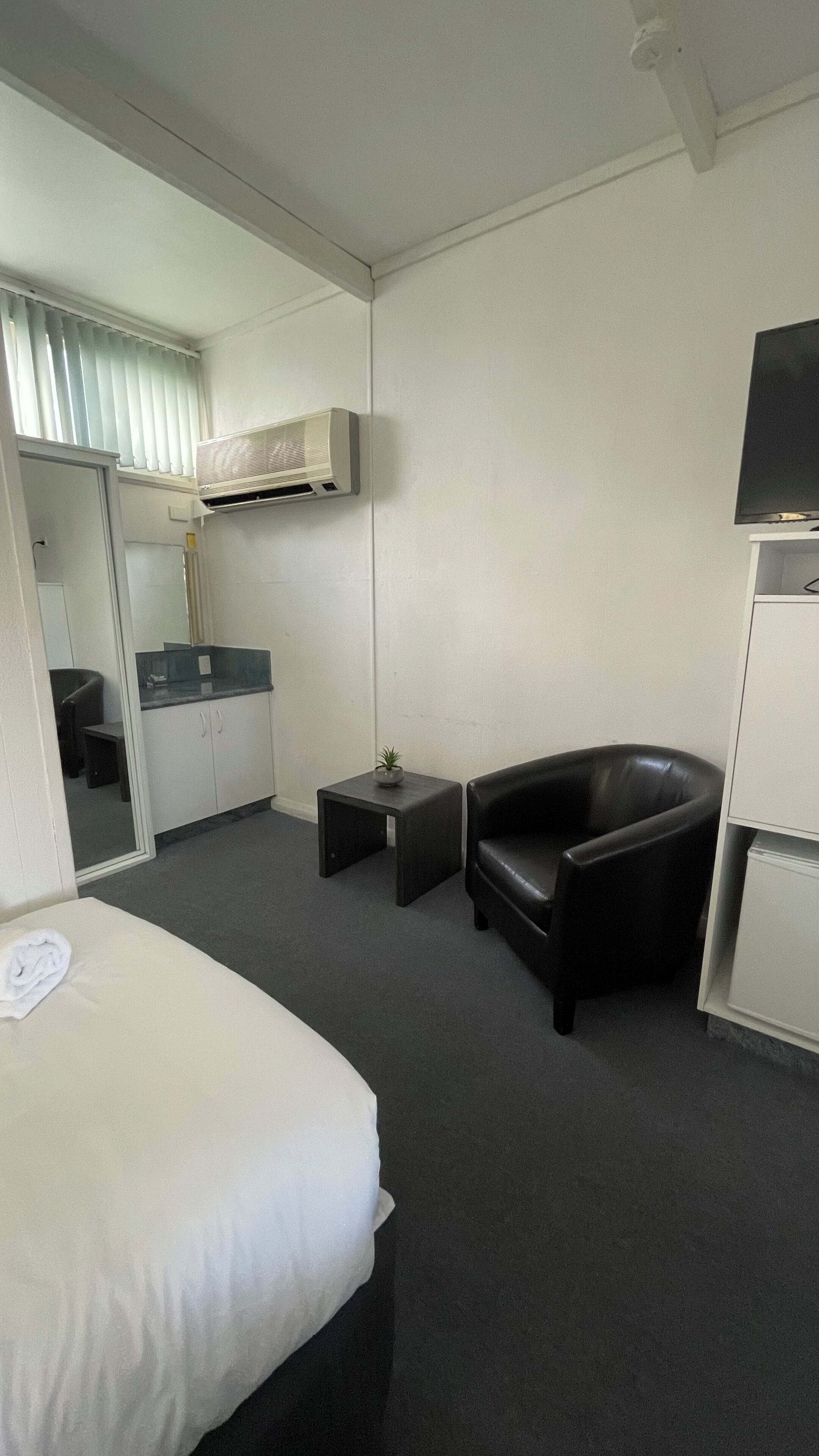 Habitación de hotel con una cama, un sillón de cuero negro, una mesita con una planta, una kitchenette y un televisor montado en la pared.