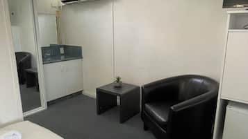 Une chambre d’hôtel avec un lit, un fauteuil en cuir noir, une petite table avec une plante, une kitchenette et un téléviseur fixé au mur.