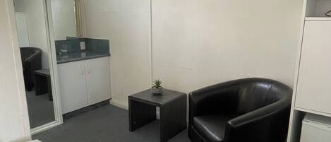 Habitación de hotel con una cama, un sillón de cuero negro, una mesita con una planta, una kitchenette y un televisor montado en la pared.