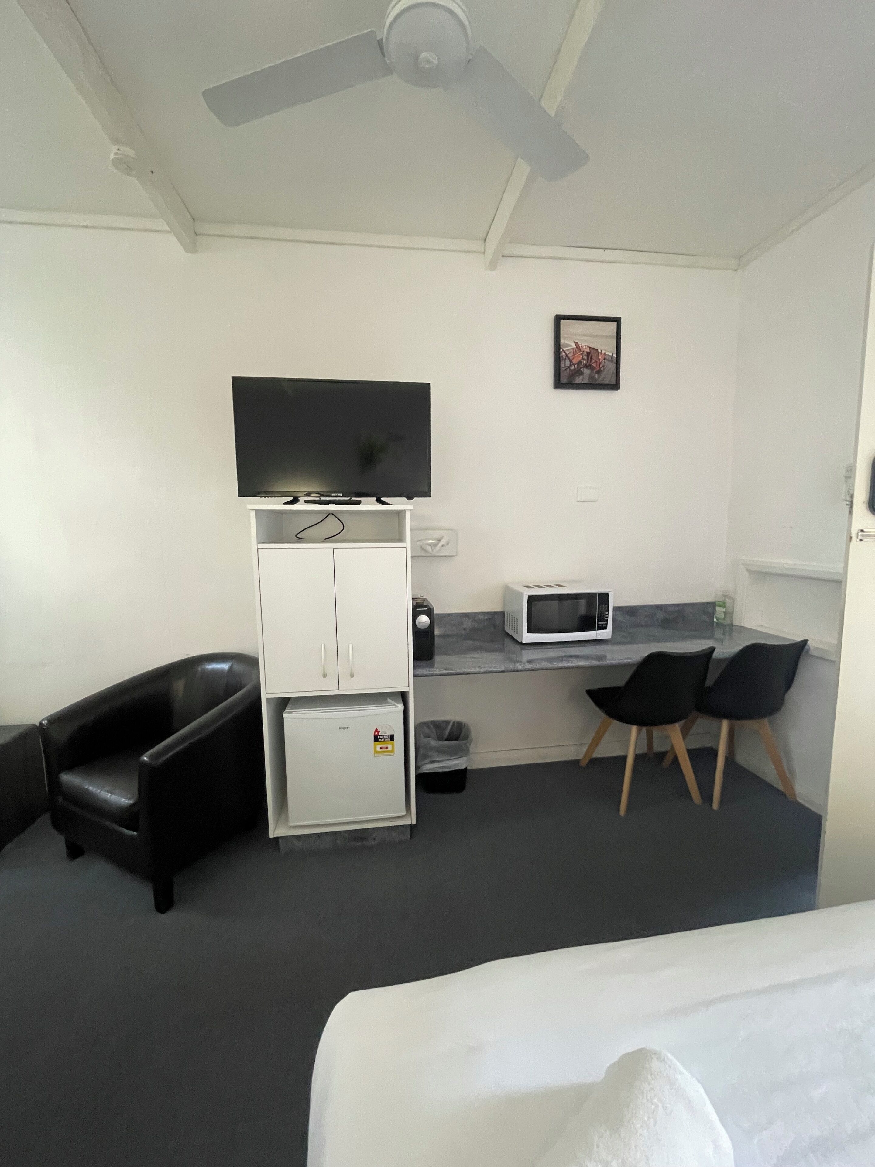 A hotel room with a bed, a desk with a microwave, a television, a refrigerator, a trash bin, and two chairs.