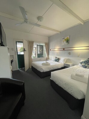 A hotel room with two beds, a ceiling fan, a window with curtains, a bedside table, and a wall-mounted light fixture. - The Tree Motel (Narooma)
