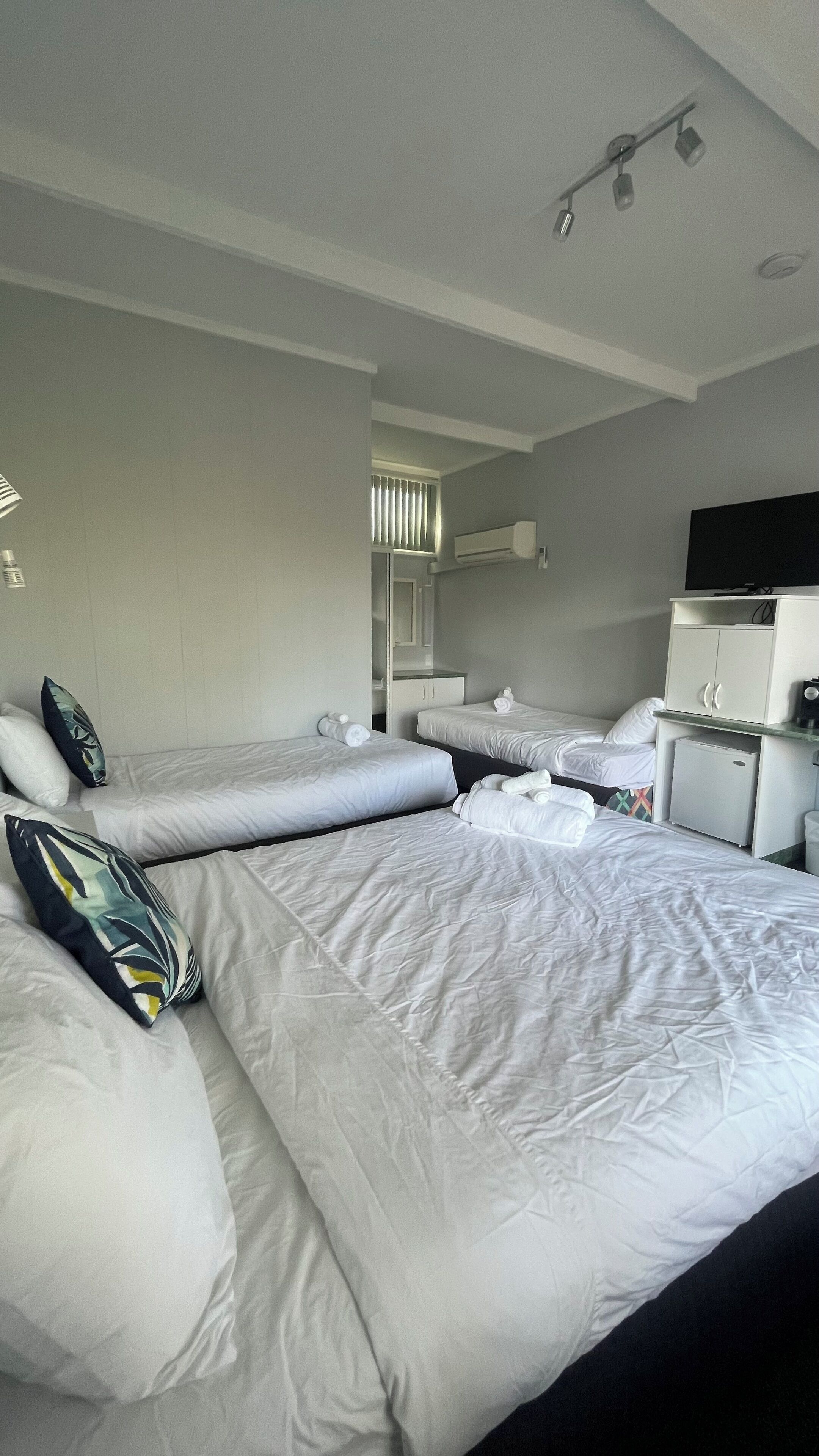 A hotel room with two single beds, a small desk with a refrigerator, a television, and a window with blinds.