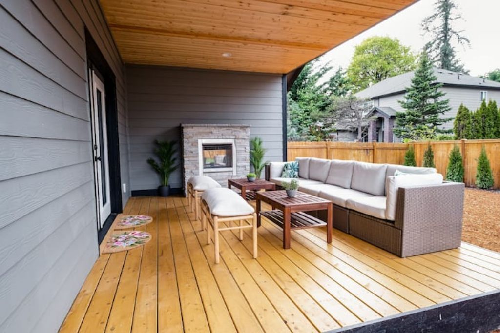 A covered patio with a wooden deck, a stone fireplace, a sofa, and a dining table.