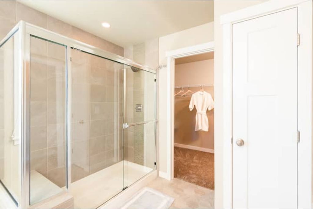 A modern bathroom with a glass-enclosed shower, a walk-in closet with hanging robes, and a white door.