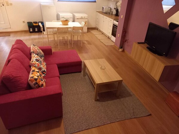 A compact living space with a red sofa, a wooden coffee table, a TV on a wooden stand, and a kitchenette in the background.