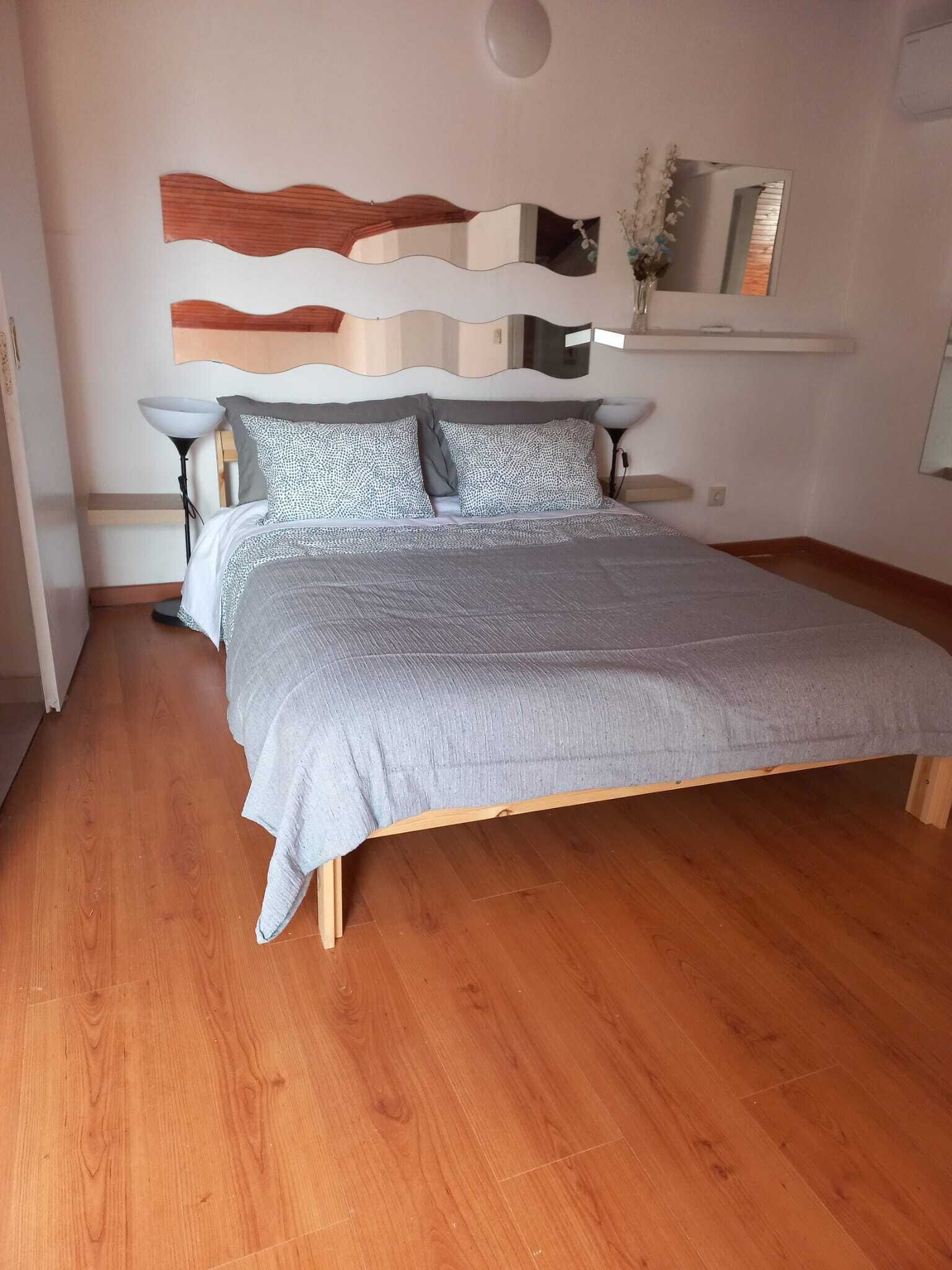 A bedroom with a wooden floor, a bed with grey bedding, a wall-mounted mirror, and a bedside table with a lamp.