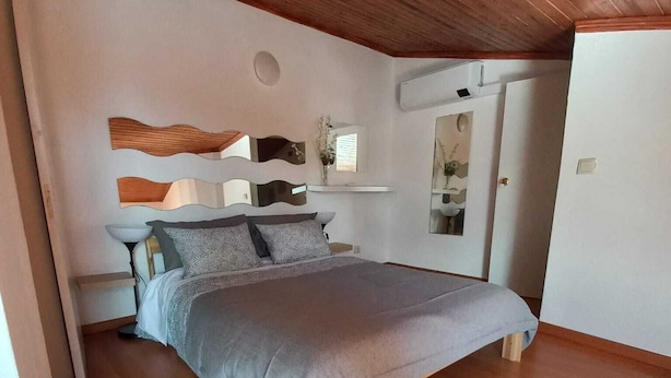 A bedroom with a wooden ceiling, a bed with grey bedding, a nightstand, and a wall-mounted mirror.