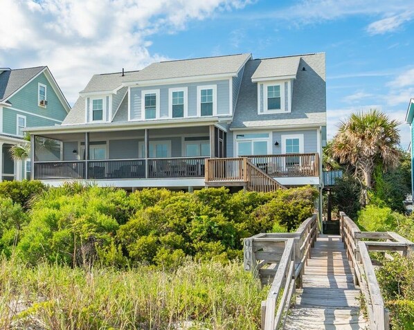 A coastal house with a wooden deck leading to a balcony, surrounded by lush greenery.