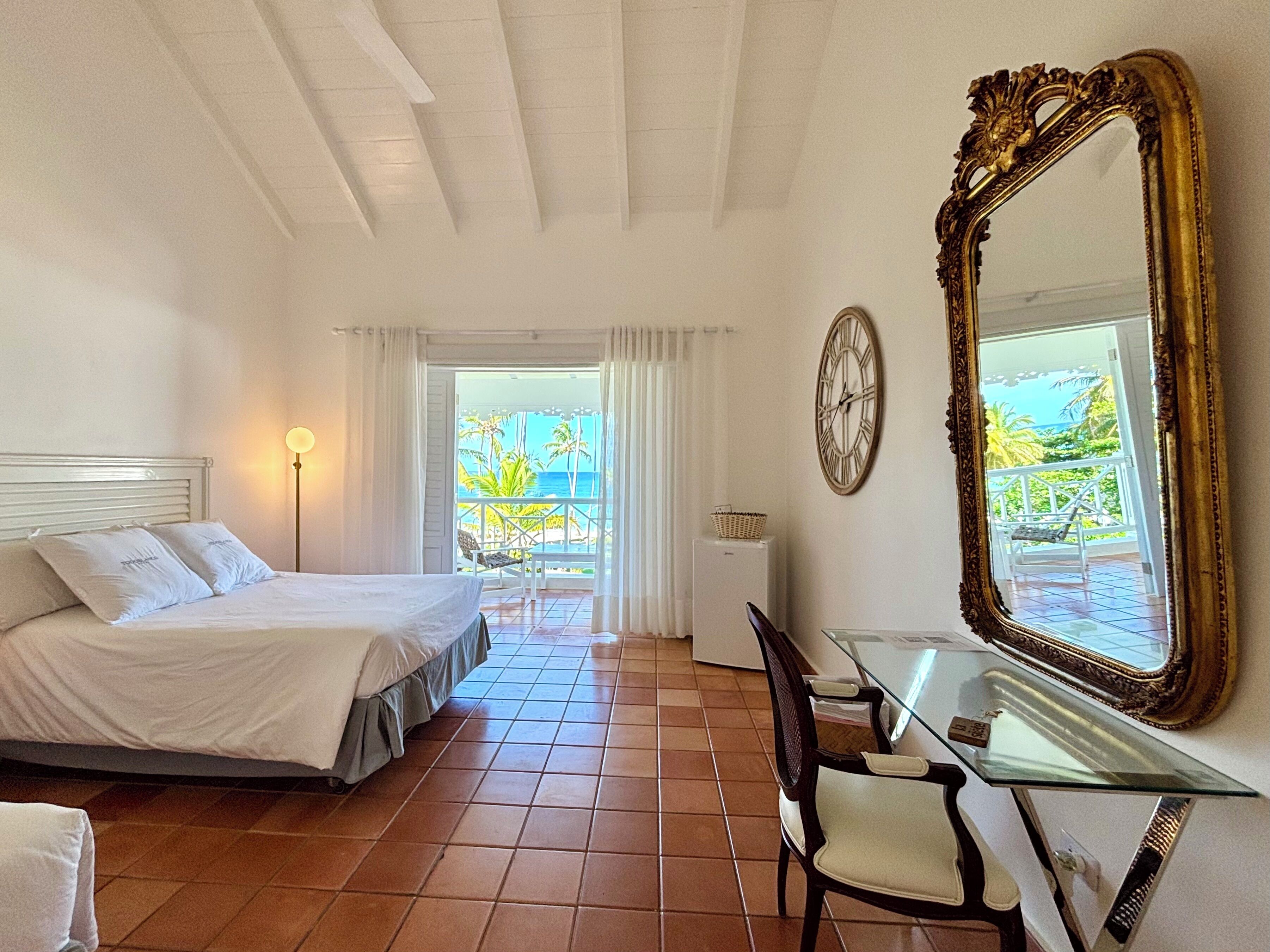 A bedroom with a bed, a desk, a chair, a large mirror, a clock, and a view of the ocean through a window.