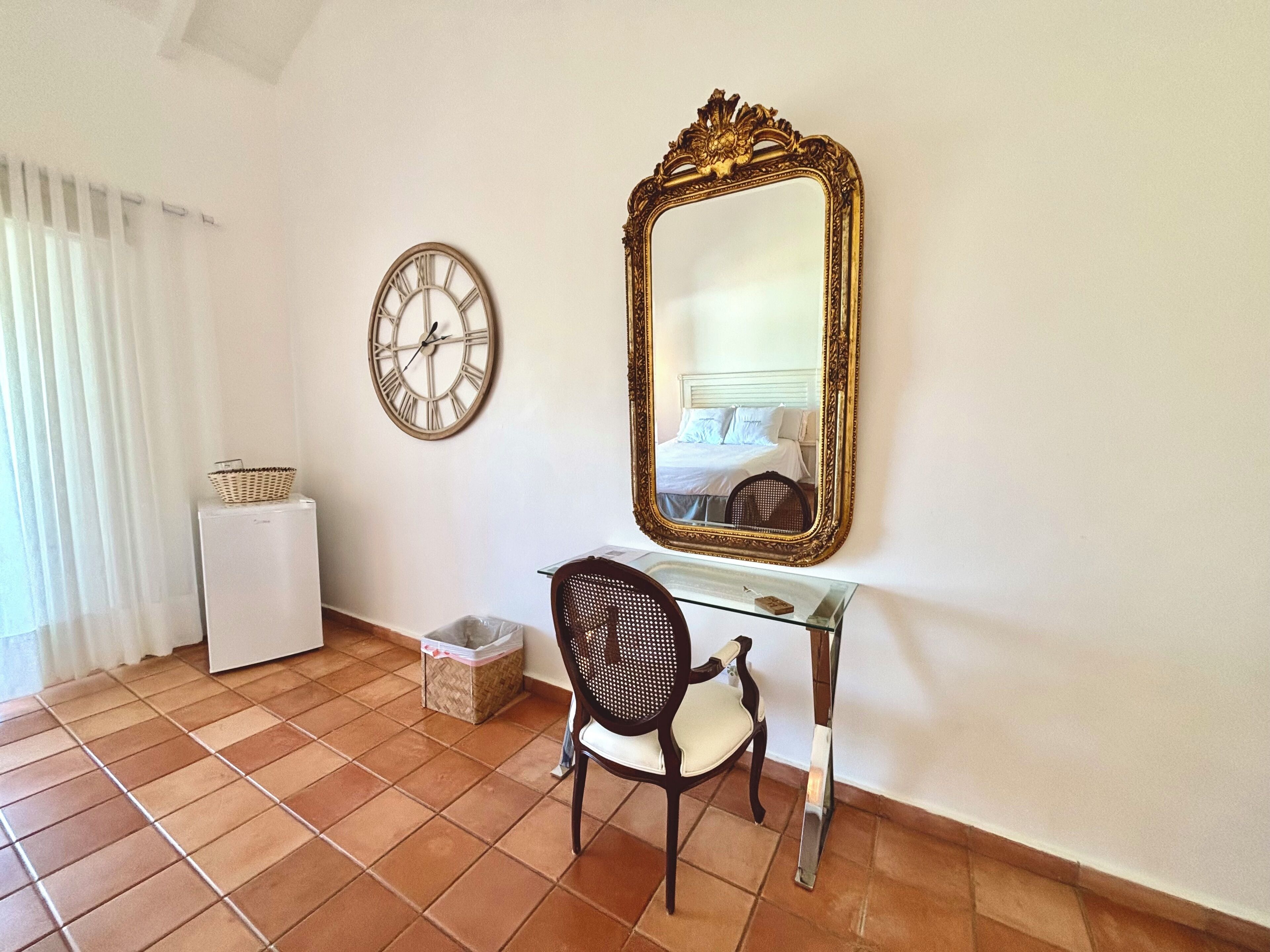 A room with a bed, a large mirror, a clock, a chair, and a small table.