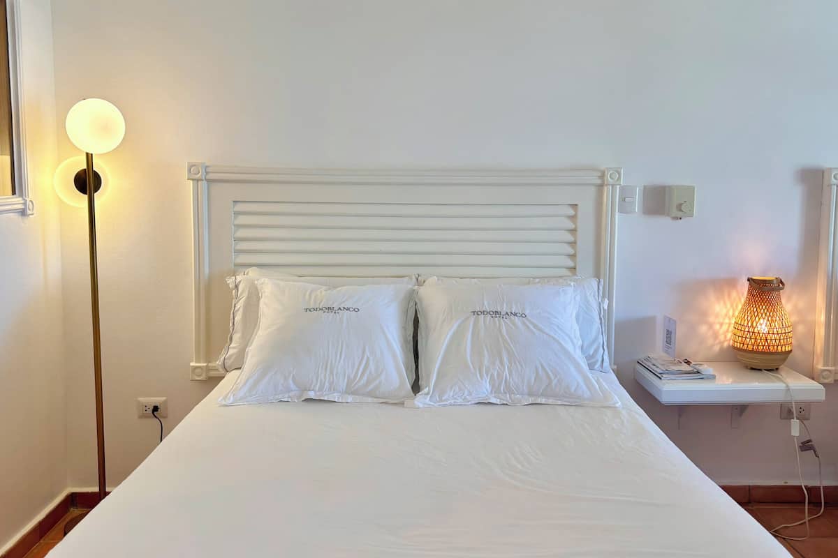 Una cama bien hecha con ropa blanca y dos almohadas, una mesita de noche con lámpara y un aplique de pared.