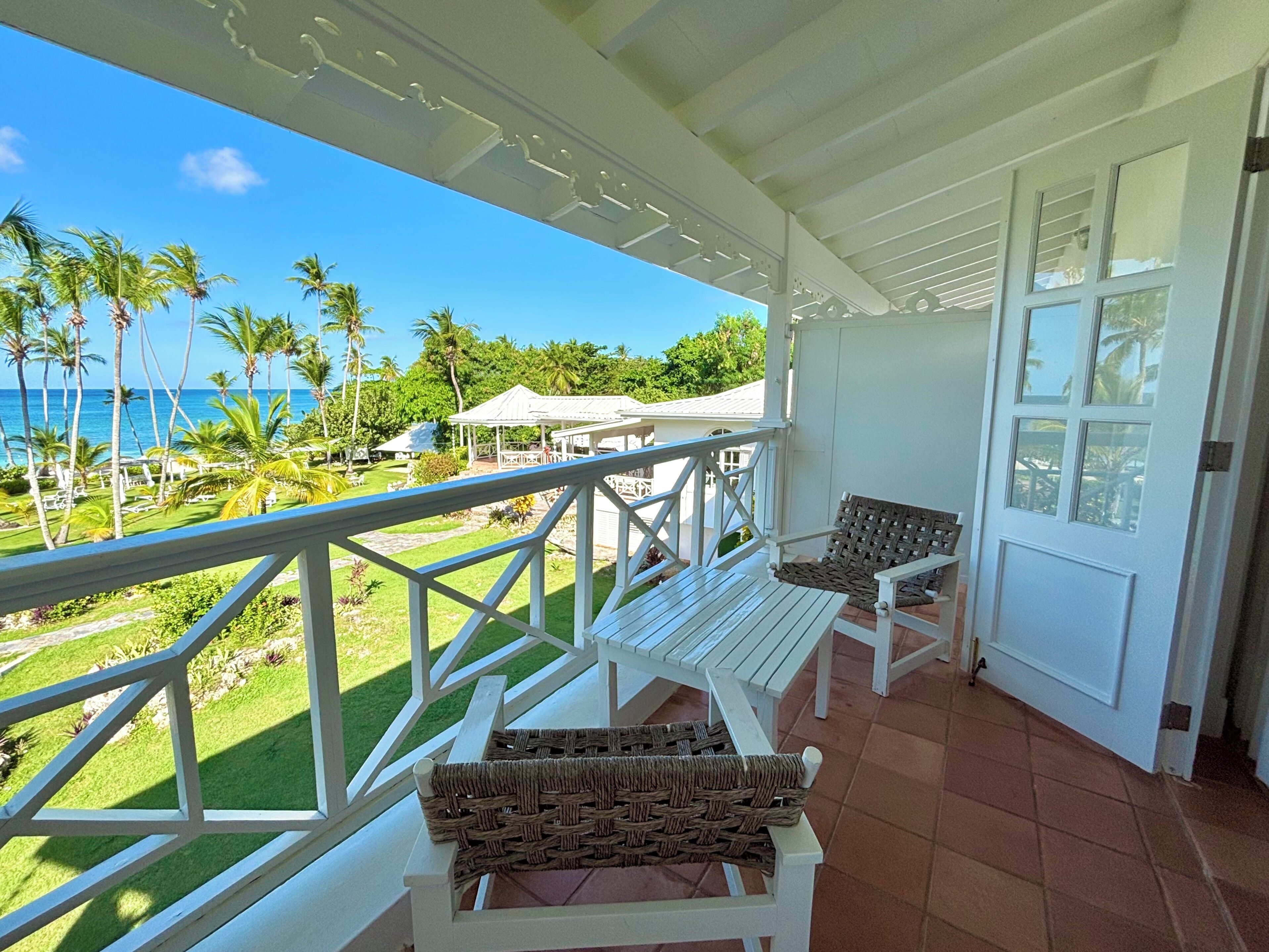 A balcony with white railings, wicker chairs, and a view of the ocean and palm trees.