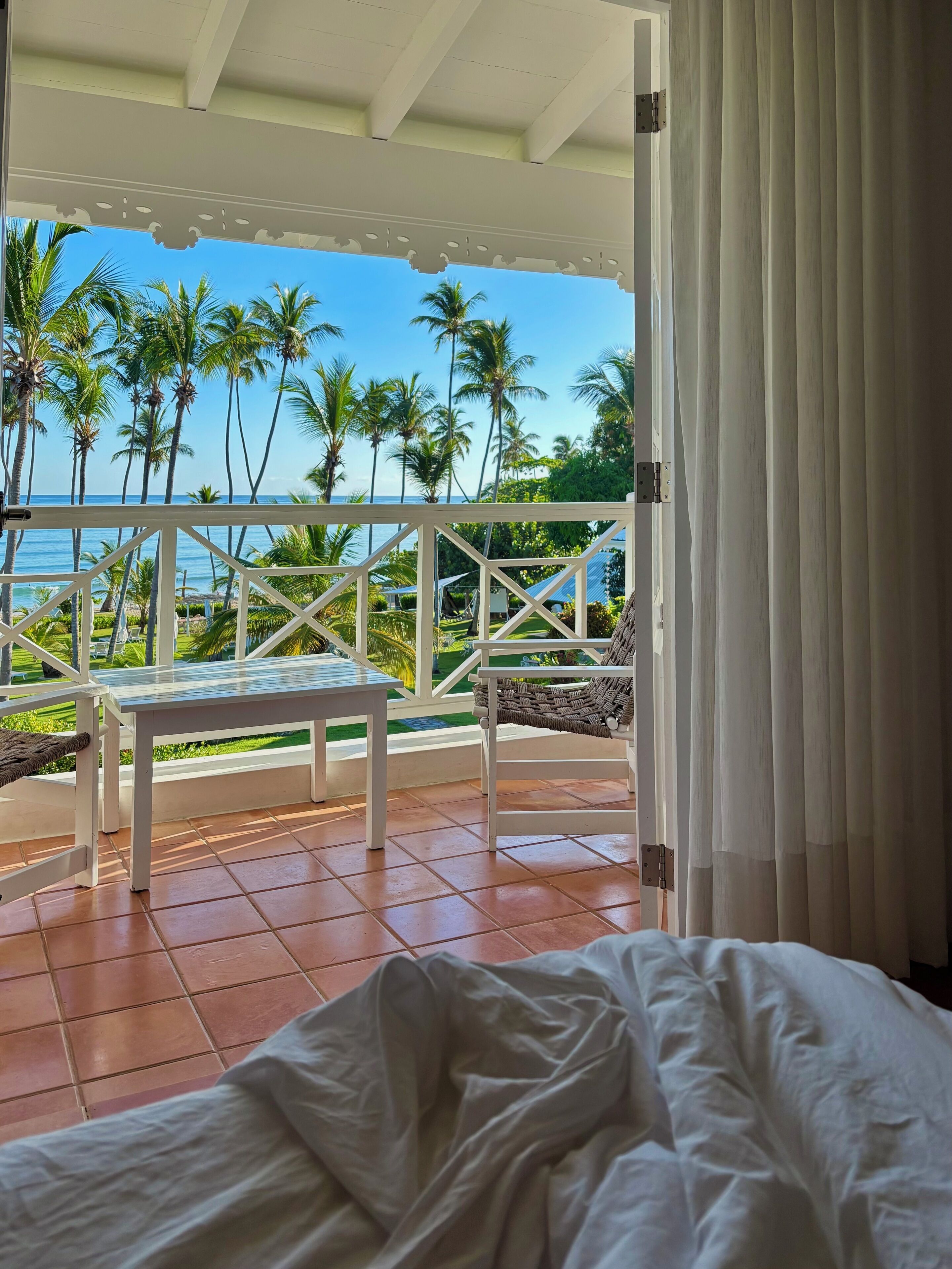 A balcony with a view of the ocean and palm trees, a white bed, and a table.