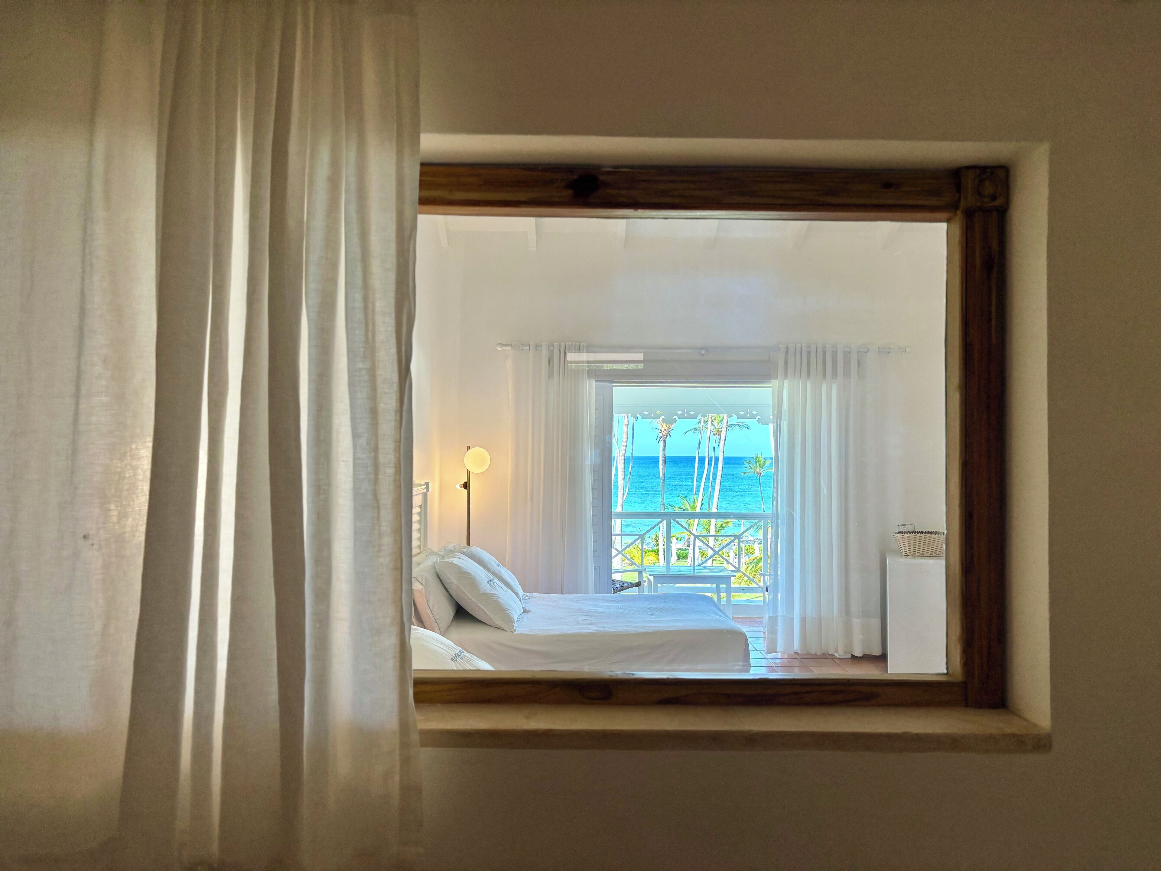 A bedroom with a view of the sea through a window, a bed, a nightstand, and a lamp.