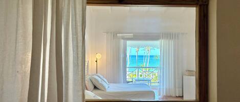 A bedroom with a view of the sea through a window, a bed, a nightstand, and a lamp.