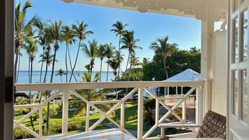 A covered outdoor area with a white bench and wicker chair, overlooking a lawn with palm trees and a view of the sea.