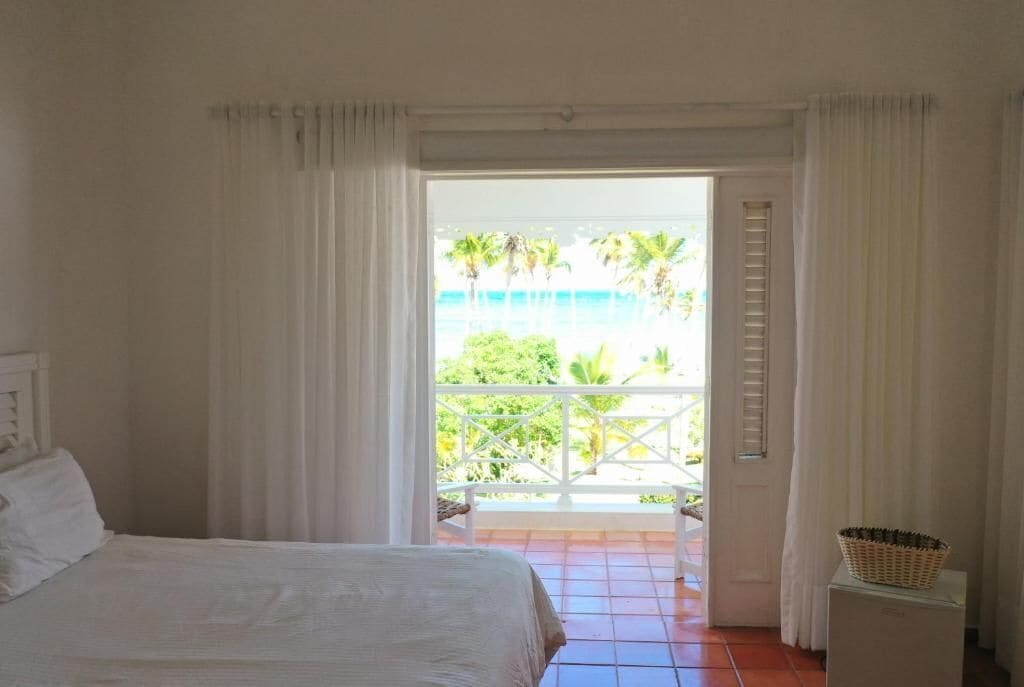 A bedroom with a bed, white curtains, a balcony with a view of palm trees and the ocean.