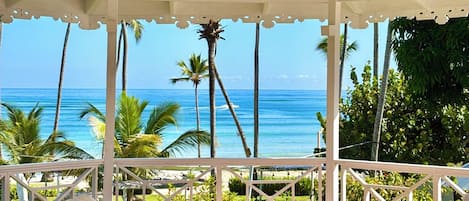 A covered outdoor seating area with white chairs and a view of the ocean and palm trees.