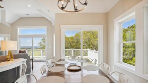 A spacious dining area with a wooden table, white chairs, and large windows offering views of the outdoors. - Sound Views, Hot Tub, Village at Ocean Hill, Community Pools (Corolla)
