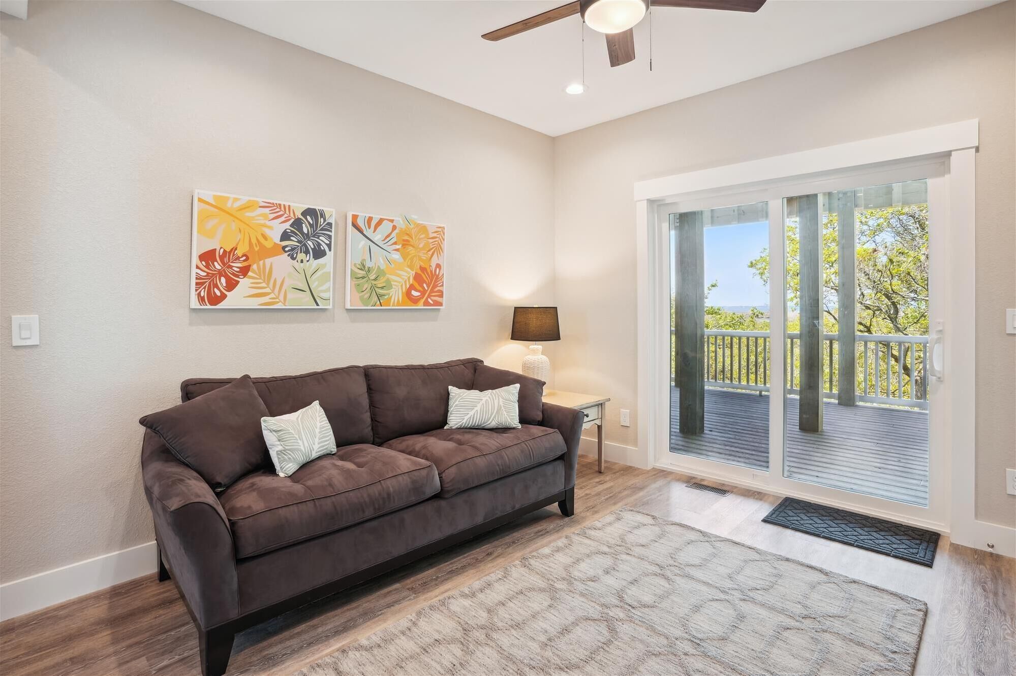 A living room with a brown sofa, two wall-mounted artworks, a side table with a lamp, and a view of the outdoors through a sliding glass door.