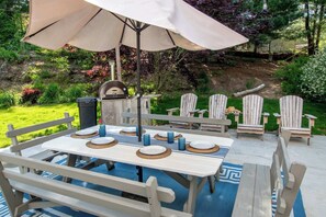 An outdoor dining area with a table set for four, surrounded by Adirondack chairs, under a large umbrella.