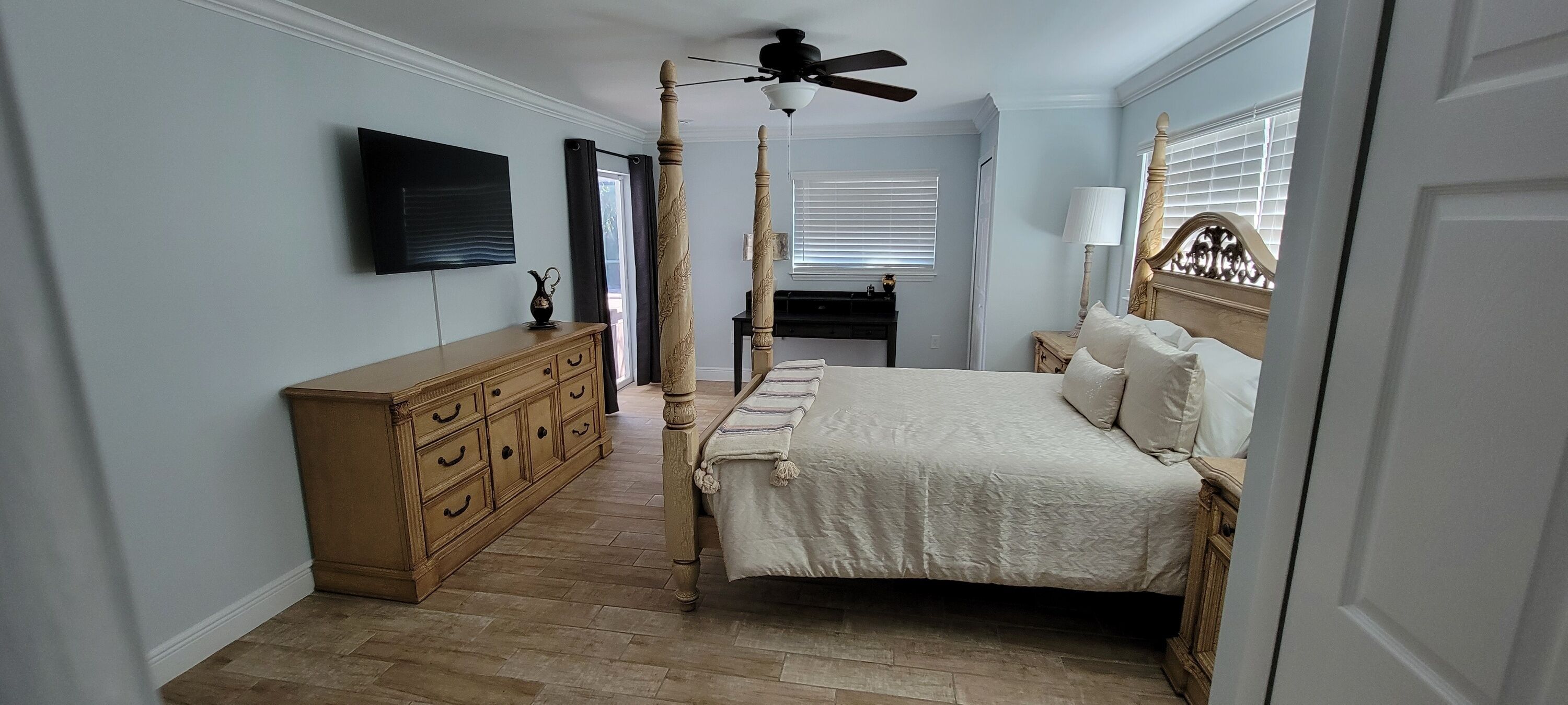 A bedroom with a four-poster bed, a wooden dresser, a television mounted on the wall, and a ceiling fan.