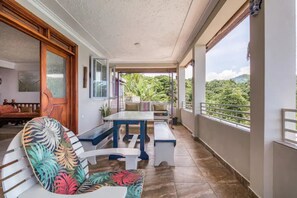A balcony with a table and benches, a chair with a floral pattern, and a view of greenery. - Braata @ Little Bay (Ocean View Villa) (Port Maria)