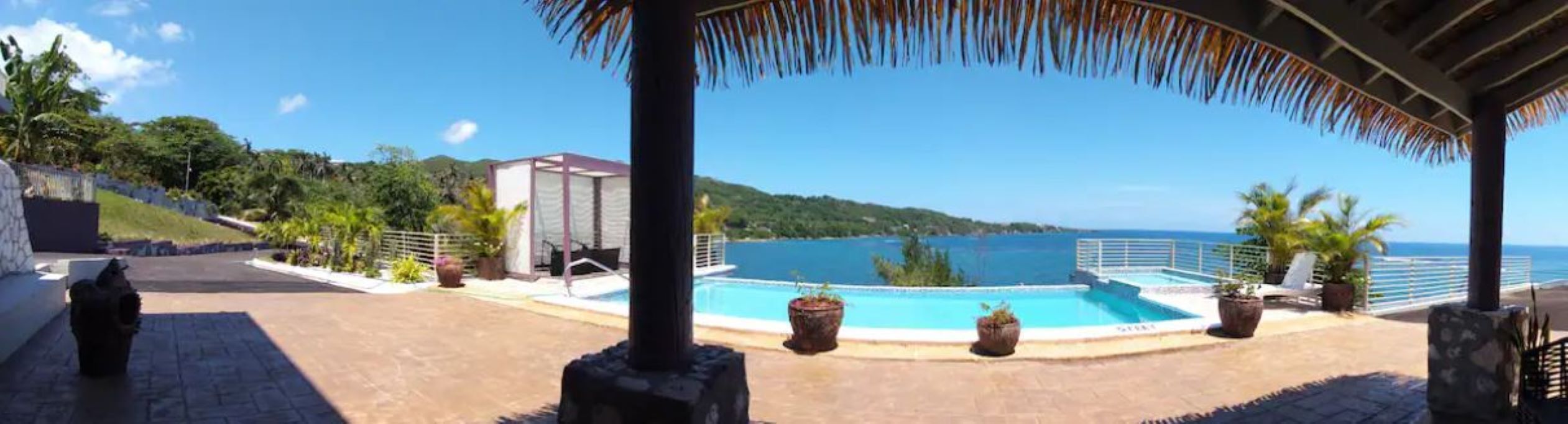 A pool area with a view of the sea, a covered seating area, and potted plants.