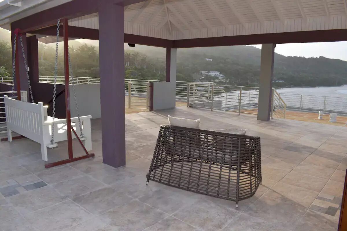 Un espace extérieur abrité comprenant une balançoire, un fauteuil en osier et une vue sur un plan d’eau et des collines.